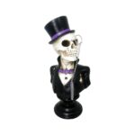 Miiniatura de busto de caveira vestida com cartola e smoking pretos, gravata borboleta roxa e camisa branca, monóculo dourado. A caveira olha para o lado direito e tem os braços cruzados para trás do tronco. Disponível para locação.